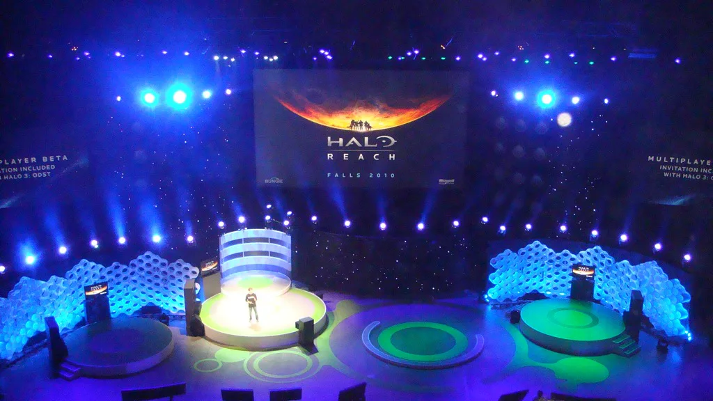 Palco de evento de games iluminado em azul e verde, com estruturas geométricas de LED e plataformas circulares, exibindo logo e arte de Halo Reach em grande tela central