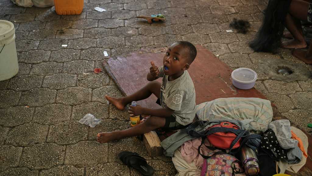 Criança sentada no chão com seus pertences em uma rua após chegada de deportados no Haiti