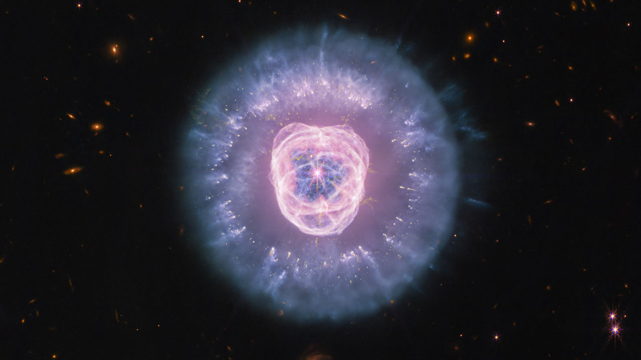 Imagem de telescópio espacial mostra nebulosa planetária com núcleo rosa-avermelhado circundado por halos de gás ionizado em tons de azul e violeta, cercada
