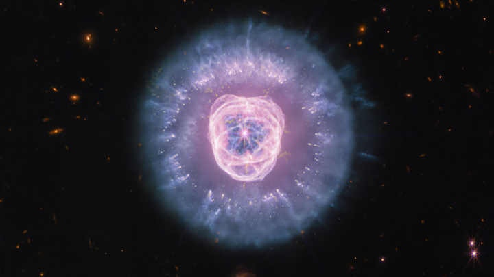 Imagem de telescópio espacial mostra nebulosa planetária com núcleo rosa-avermelhado circundado por halos de gás ionizado em tons de azul e violeta, cercada