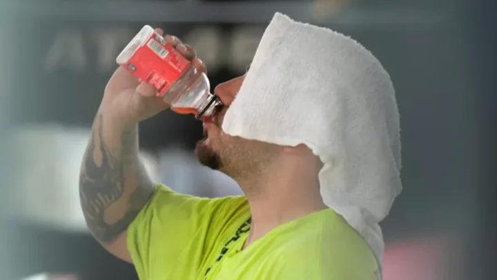 Homem de camiseta amarela fluorescente bebe água de garrafa enquanto se protege do calor com toalha sobre a cabeça