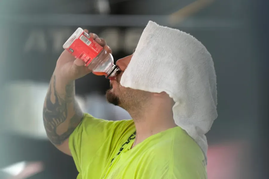 Homem de camiseta amarela fluorescente bebe água de garrafa enquanto se protege do calor com toalha sobre a cabeça