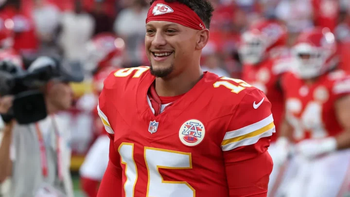 Jogador de futebol americano com uniforme vermelho e branco sorri durante partida no estádio, com torcedores ao fundo