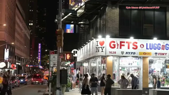 Rua movimentada à noite em Times Square com pedestres caminhando em frente a loja de souvenires e presentes, enquanto veículos circulam e edifícios iluminados aparecem ao fundo