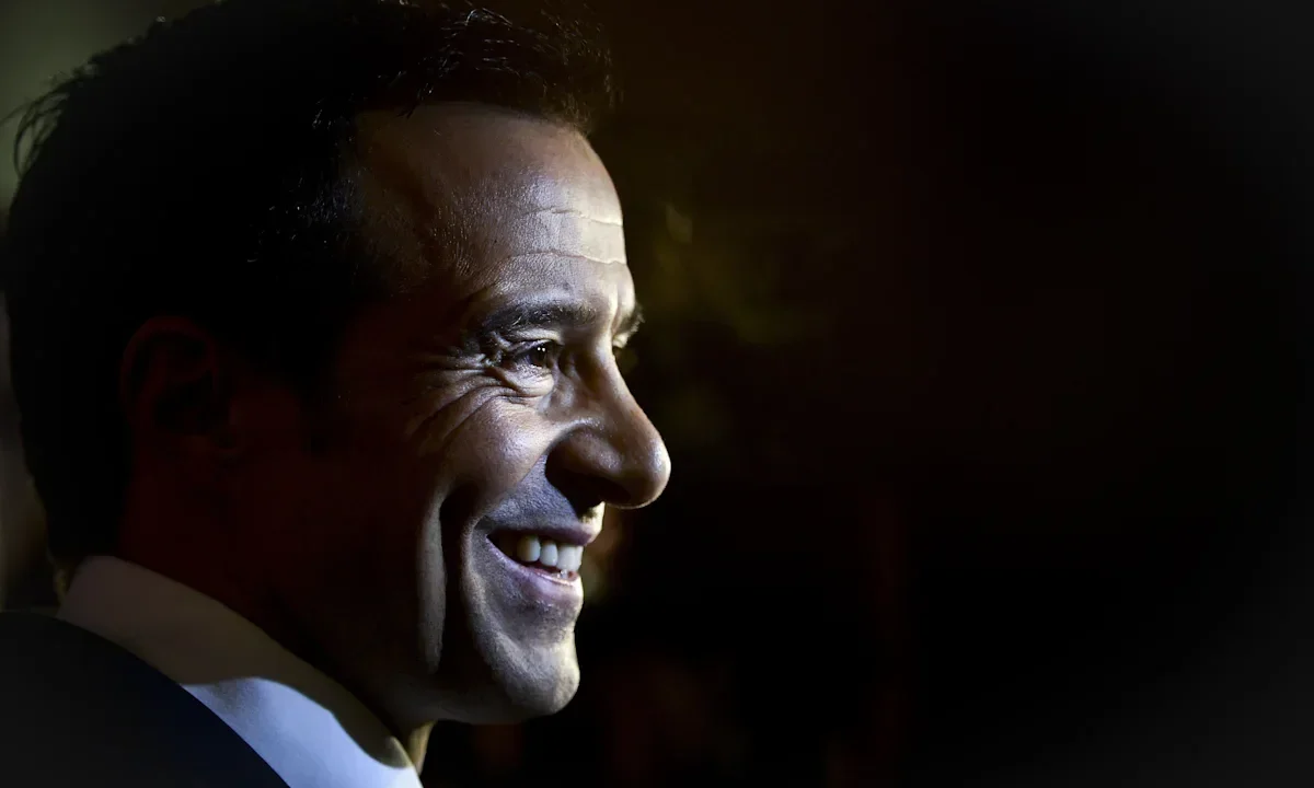 Homem de perfil, sorrindo, iluminado contra fundo escuro, usando camisa e gravata azul