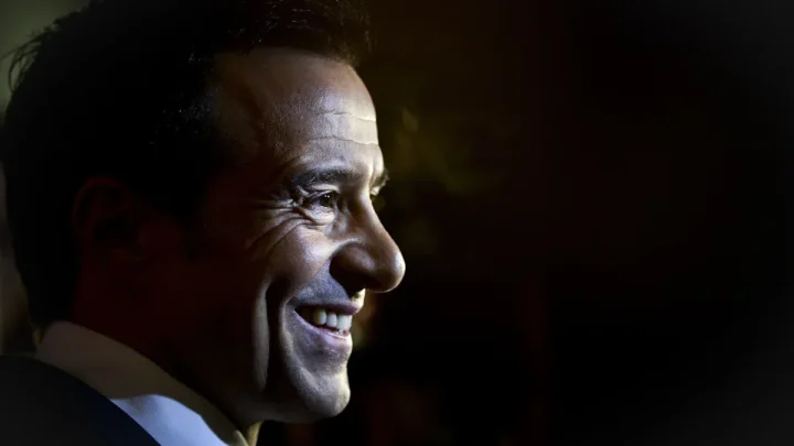 Homem de perfil, sorrindo, iluminado contra fundo escuro, usando camisa e gravata azul