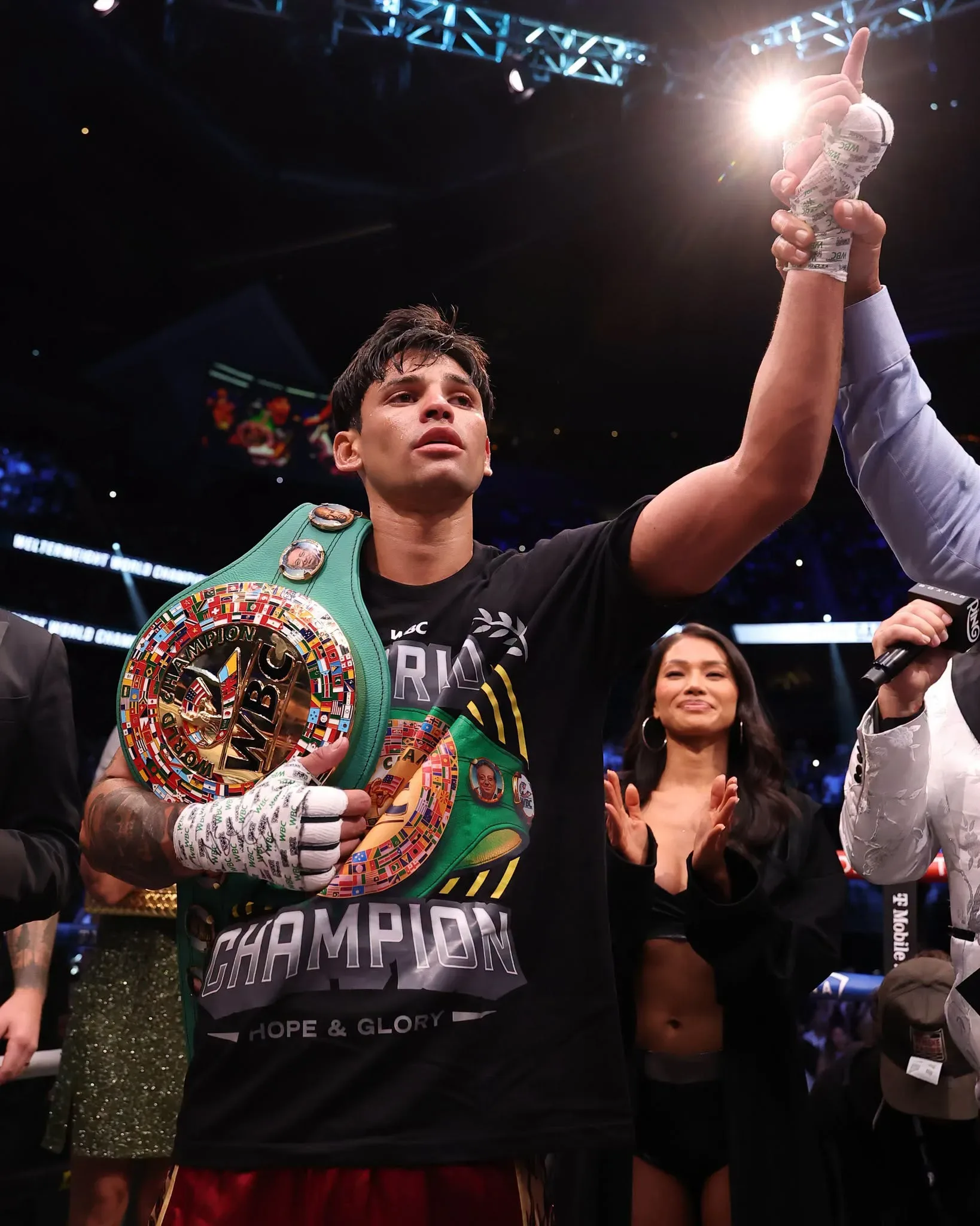 Boxeador celebra vitória no ringue com cinturão de campeão na cintura e troféu levantado ao alto, ao lado de equipe e público em arena de Las Vegas