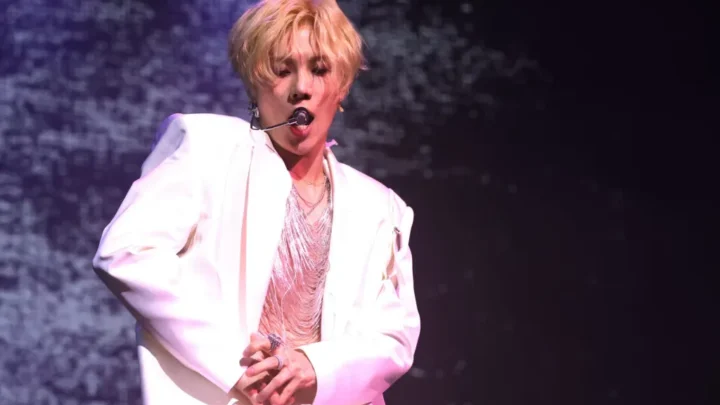 Artista com cabelo loiro curto e blazer branco se apresenta no palco com microfone headset durante festival de música