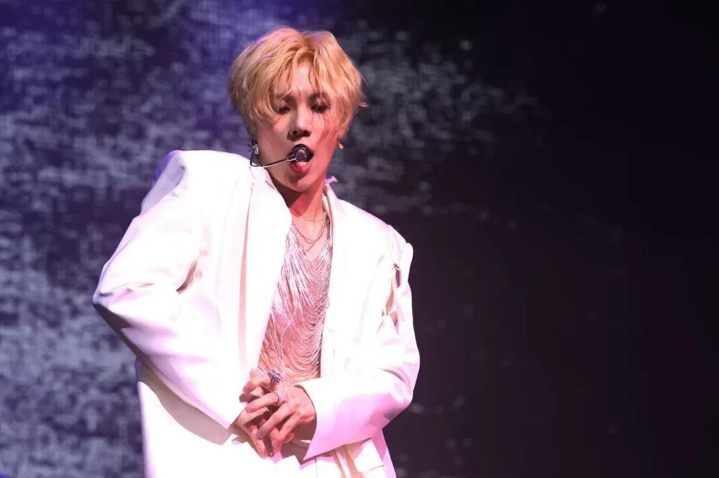 Artista com cabelo loiro curto e blazer branco se apresenta no palco com microfone headset durante festival de música