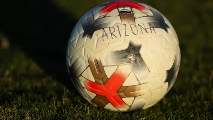 Bola de futebol com logotipo da Arizona sobre grama em estádio, durante partida entre universidades