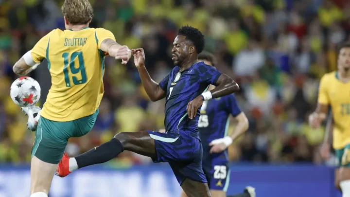 Jogador de uniforme azul disputa a bola com atleta de amarelo durante partida de futebol em estádio com público ao fundo