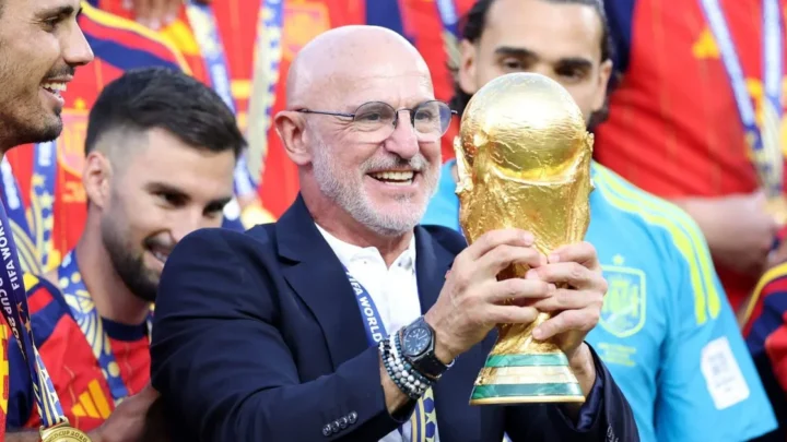 Homem de terno e óculos segura a Taça do Mundo de Futebol durante celebração com jogadores em uniforme vermelho e azul ao fundo