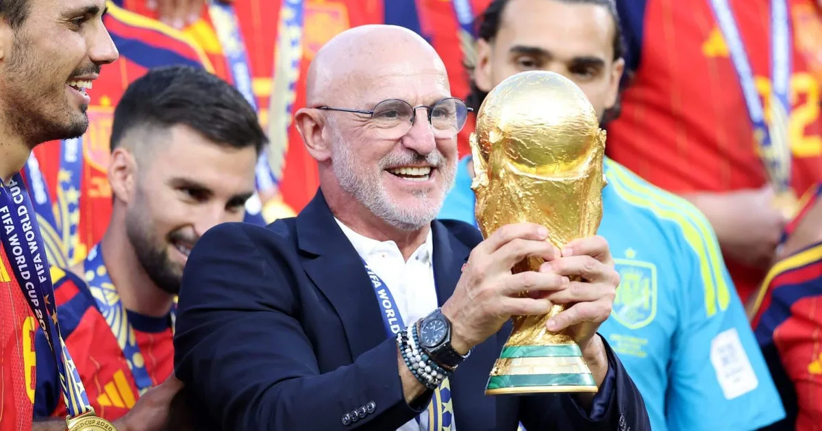 Homem de terno e óculos segura a Taça do Mundo de Futebol durante celebração com jogadores em uniforme vermelho e azul ao fundo