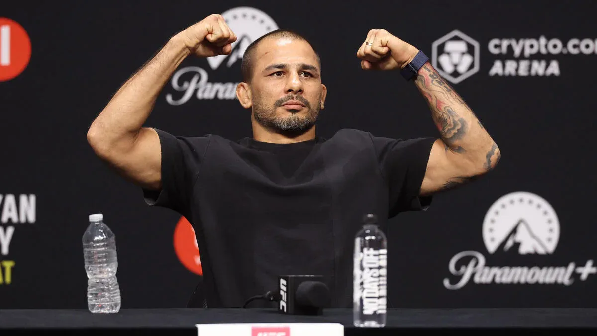 Atleta de MMA em coletiva de imprensa flexiona os músculos dos braços em gesto de confiança antes de confronto no UFC, em arena patrocinada por criptomoeda