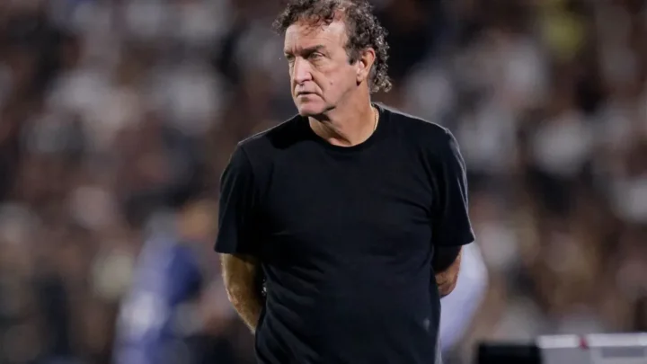 Homem de camiseta preta observa partida de futebol em estádio com torcida ao fundo