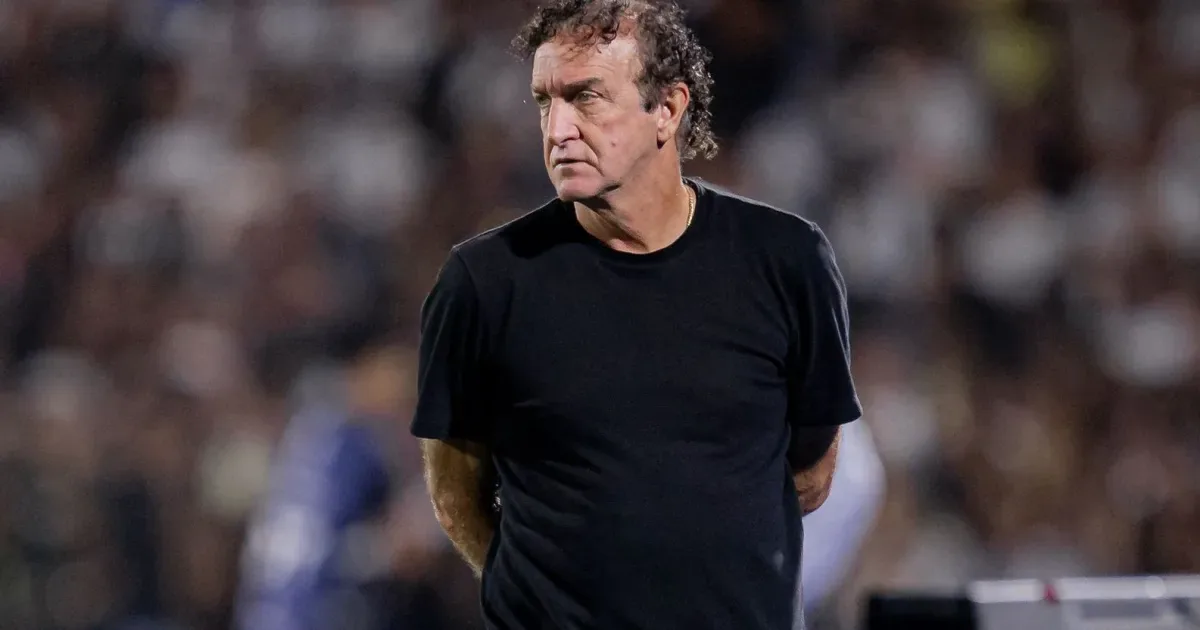 Homem de camiseta preta observa partida de futebol em estádio com torcida ao fundo