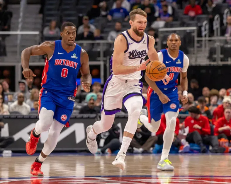 Jogador da equipe visitante em uniforme branco com detalhes em roxo conduz a bola enquanto dois adversários em azul e vermelho tentam marcá-lo durante partida da NBA