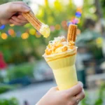 Mão segura copo com bebida amarela, calda, pipoca e biscoito de canela em parque ao ar livre