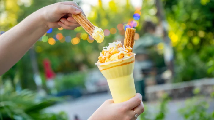 Mão segura copo com bebida amarela, calda, pipoca e biscoito de canela em parque ao ar livre