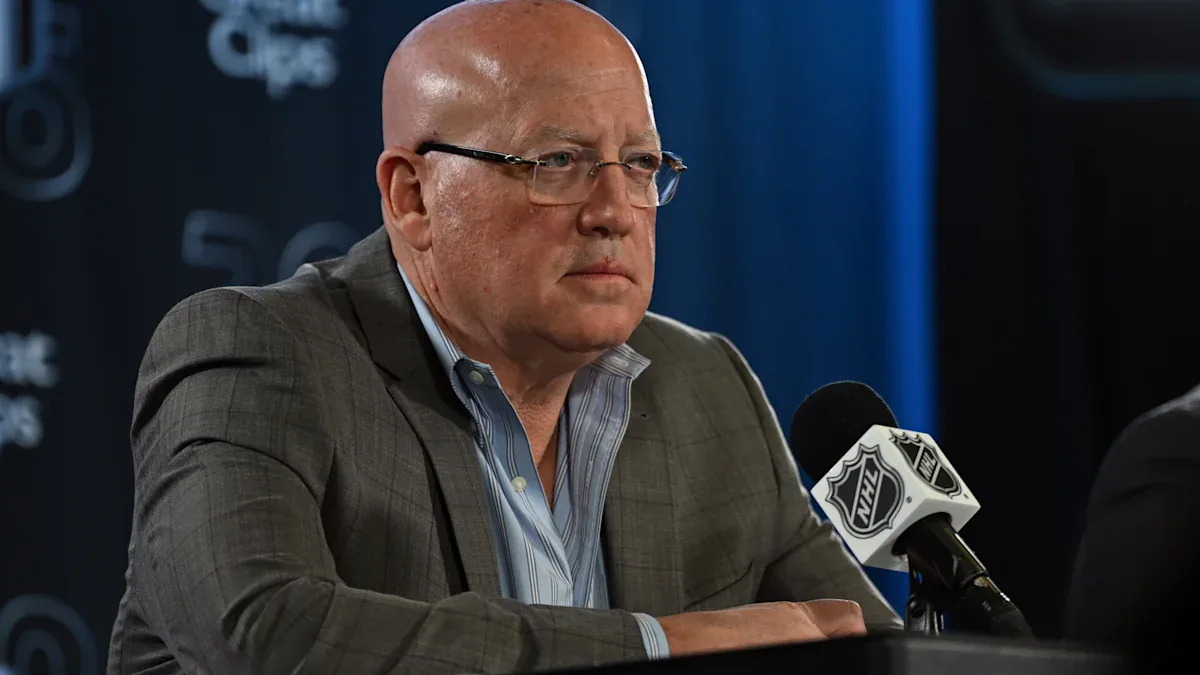 Homem de terno e óculos fala ao microfone em coletiva de imprensa durante evento da NHL, com backdrop azul ao fundo
