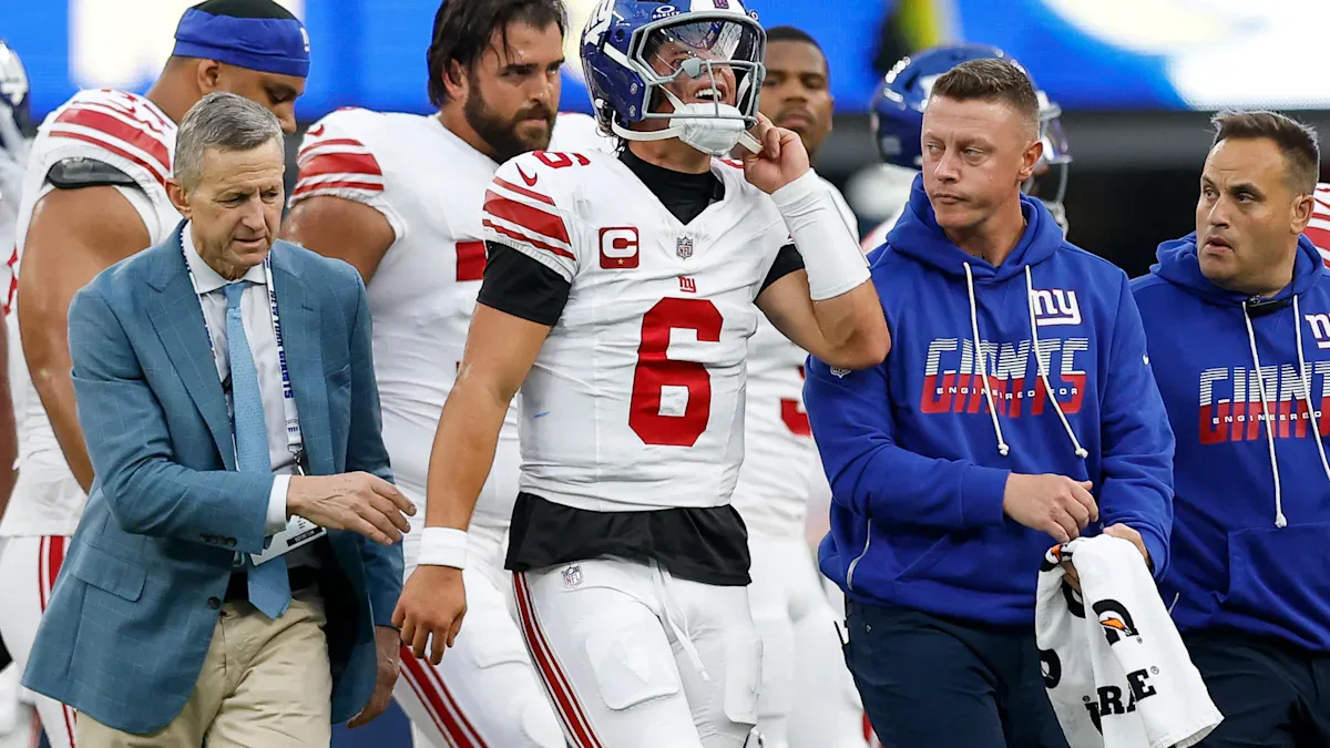 Jogador de futebol americano em uniforme branco e vermelho conversa com membros da comissão técnica durante partida