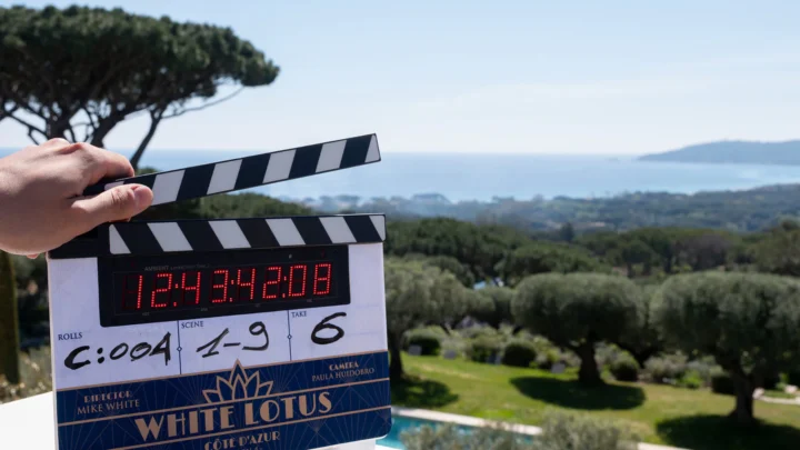 Mão segura claquete de filmagem com informações de cena durante gravação de produção audiovisual em locação com vista para costa litorânea e vegetação mediterrânea