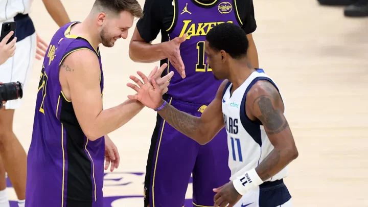 Jogadores de Lakers e Mavericks se cumprimentam durante partida na quadra, com um deles em uniforme roxo e outro em branco e azul