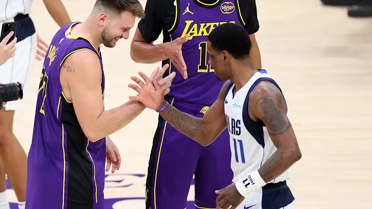 Jogadores de Lakers e Mavericks se cumprimentam durante partida na quadra, com um deles em uniforme roxo e outro em branco e azul