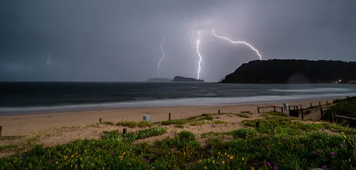 Raios iluminam o céu durante tempestade sobre praia com areia, cerca e vegetação em primeiro plano, enquanto trovoada avança sobre o oceano