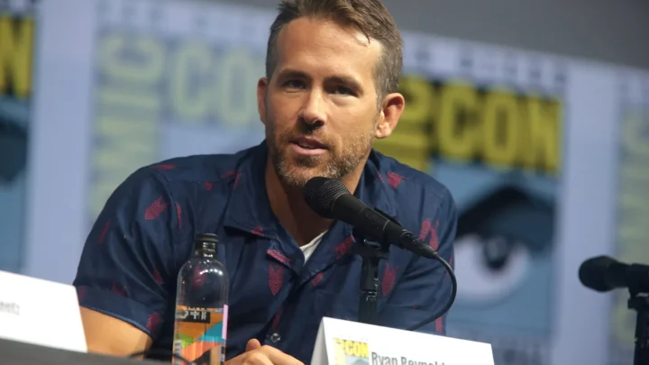 Ryan Reynolds fala em painel de evento, em frente a microfone e garrafa de água, com placa de identificação.