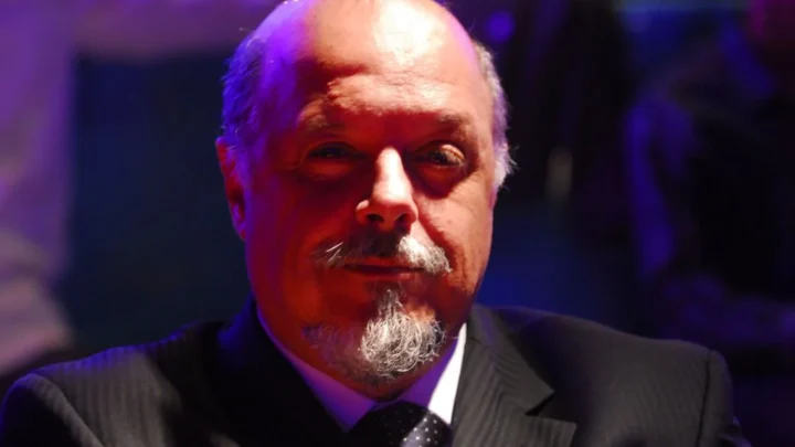 Homem de terno e gravata, com barba grisalha, em ambiente com iluminação azul e roxa