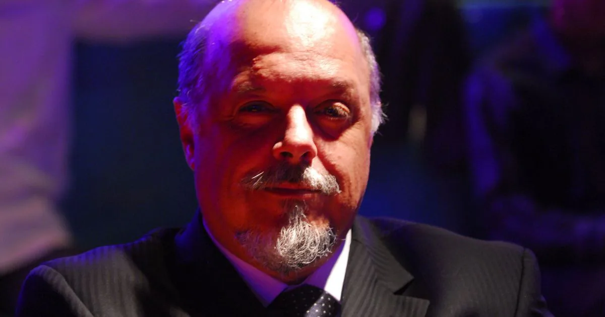 Homem de terno e gravata, com barba grisalha, em ambiente com iluminação azul e roxa