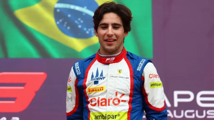 Piloto de categoria de base usando traje de corrida com patrocínios em pódio, com bandeira brasileira ao fundo