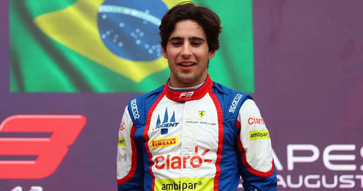 Piloto de categoria de base usando traje de corrida com patrocínios em pódio, com bandeira brasileira ao fundo