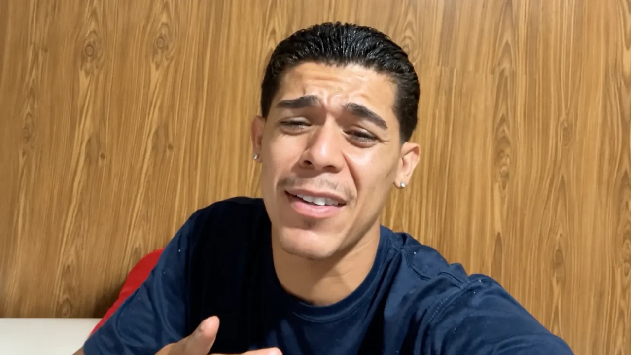 Homem de camiseta azul escura fala durante entrevista em frente a parede de madeira