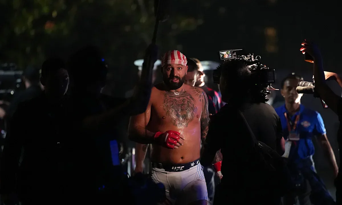 Lutador de artes marciais com luvas vermelhas e bandana listrada em branco e vermelho caminha rodeado por câmeras e equipe de produção durante evento noturno