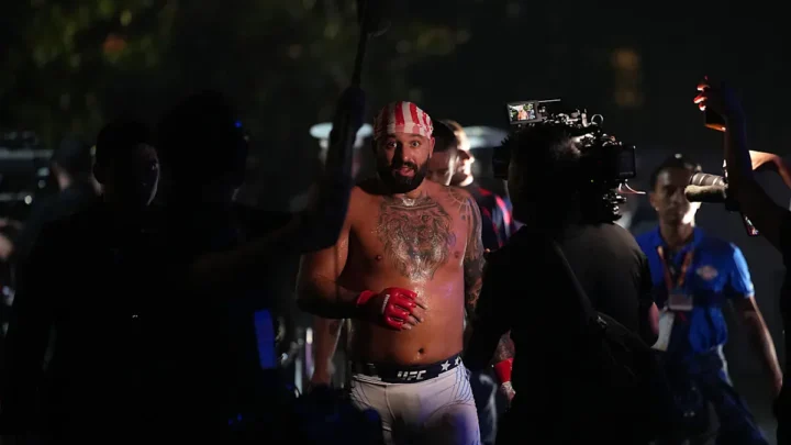 Lutador de artes marciais com luvas vermelhas e bandana listrada em branco e vermelho caminha rodeado por câmeras e equipe de produção durante evento noturno