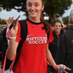 Duas jovens mulheres em uniforme de futebol de uma universidade caminham em evento ao ar livre, com uma delas segurando um microfone e sorrindo para a câmera