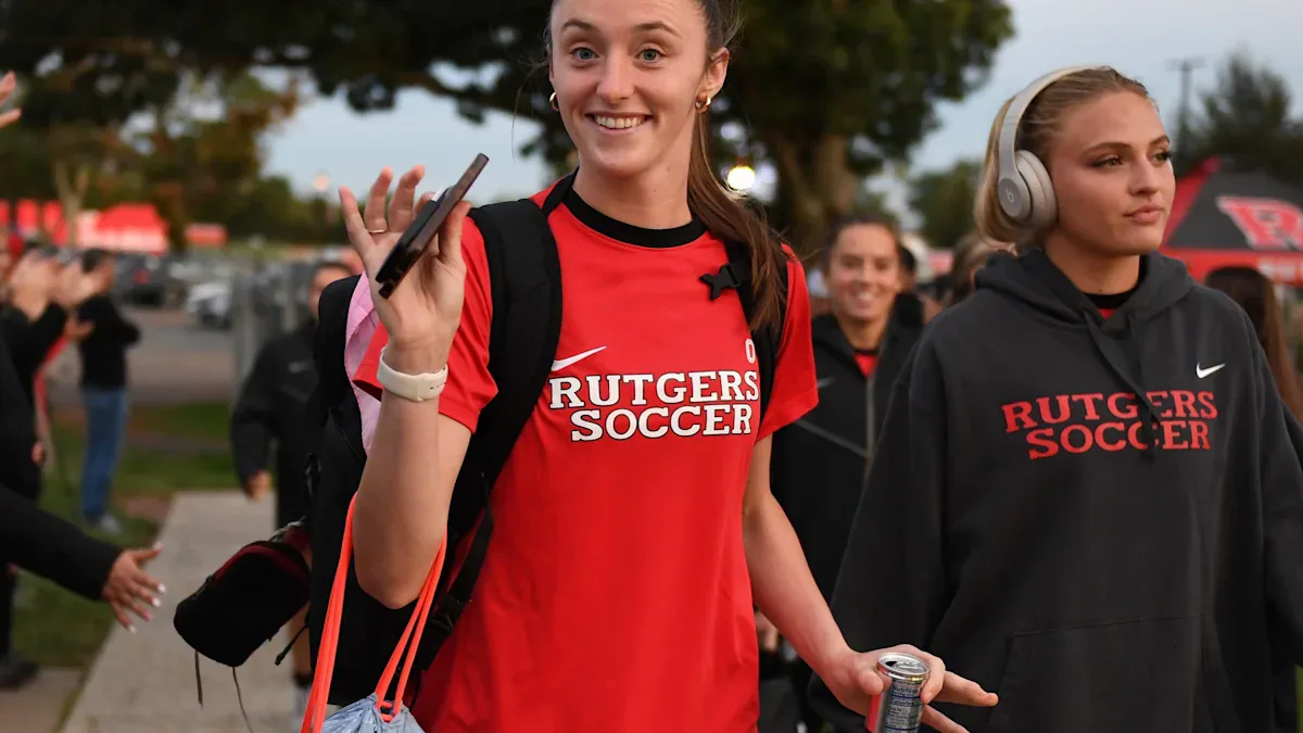 Duas jovens mulheres em uniforme de futebol de uma universidade caminham em evento ao ar livre, com uma delas segurando um microfone e sorrindo para a câmera