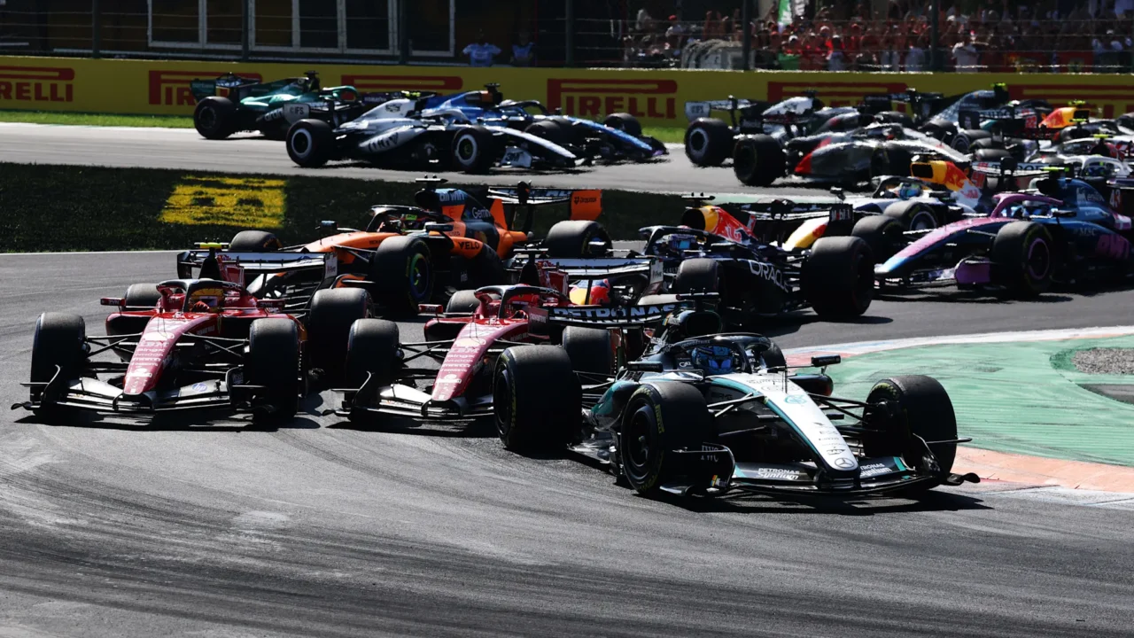 Monoposto de Fórmula 1 em disputa durante corrida em circuito urbano, com carro branco e azul à frente e monopostos vermelhos nas proximidades, enquanto público acompanha das arquibancadas