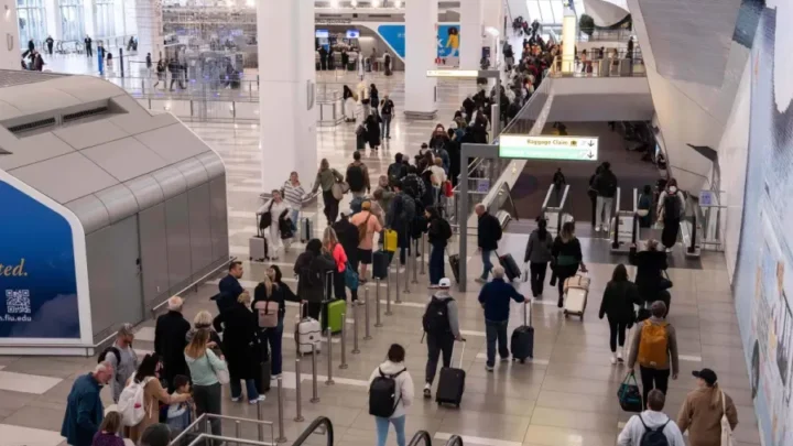 Passageiros e visitantes circulam pelo terminal de aeroporto, com filas organizadas por barreiras metálicas e pessoas com malas aguardando em diferentes áreas do saguão