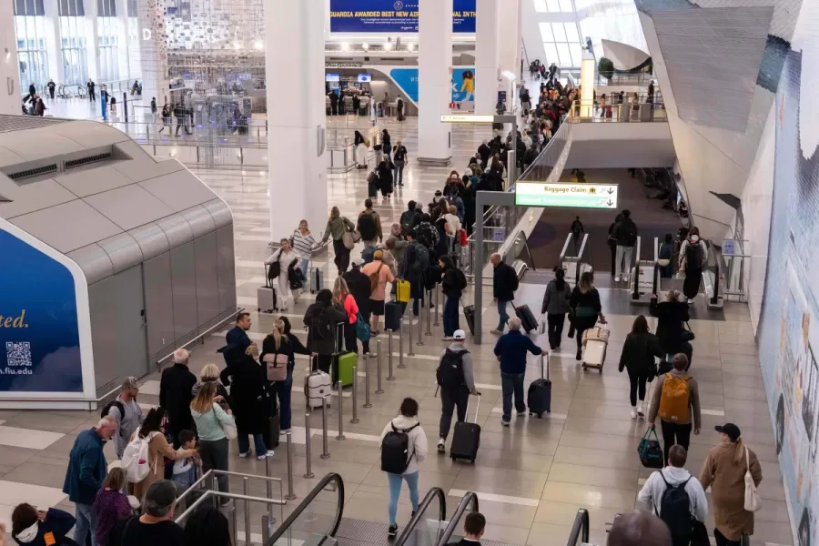 Passageiros e visitantes circulam pelo terminal de aeroporto, com filas organizadas por barreiras metálicas e pessoas com malas aguardando em diferentes áreas do saguão