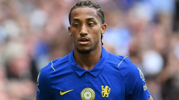 Jogador de futebol em uniforme azul com brasão de time inglês durante partida, com público ao fundo desfocado