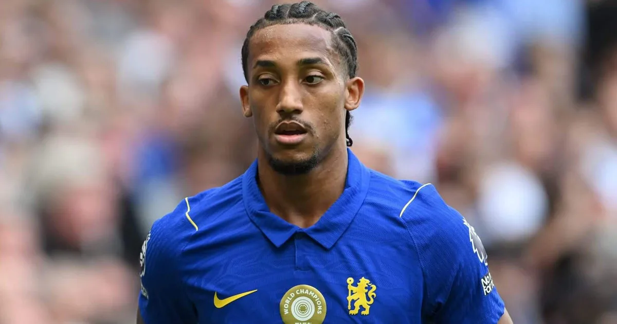 Jogador de futebol em uniforme azul com brasão de time inglês durante partida, com público ao fundo desfocado