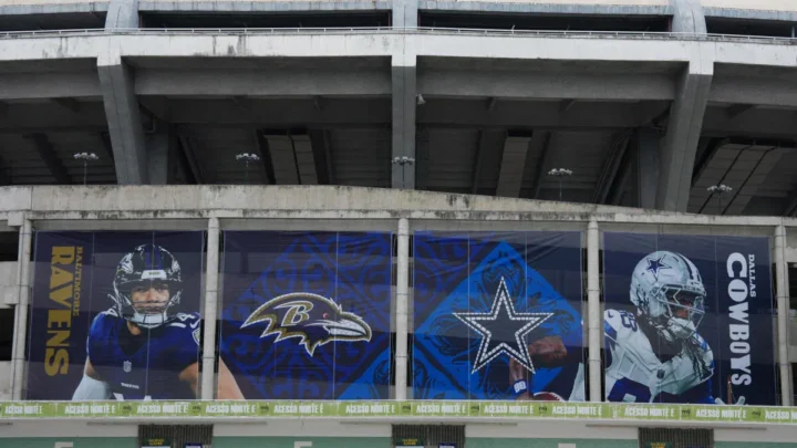 Fachada de estádio com painéis de vidro exibindo jogadores de times de futebol americano da NFL, incluindo símbolos de Ravens e Cowboys, em preparação para jogo internacional