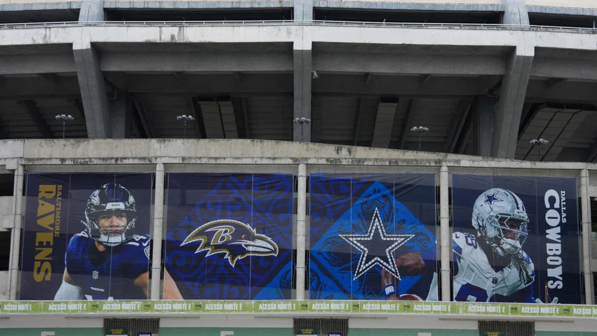 Fachada de estádio com painéis de vidro exibindo jogadores de times de futebol americano da NFL, incluindo símbolos de Ravens e Cowboys, em preparação para jogo internacional
