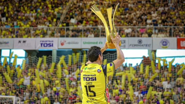 Jogador de futsal exibe troféu em estádio lotado com torcedores vestidos de amarelo
