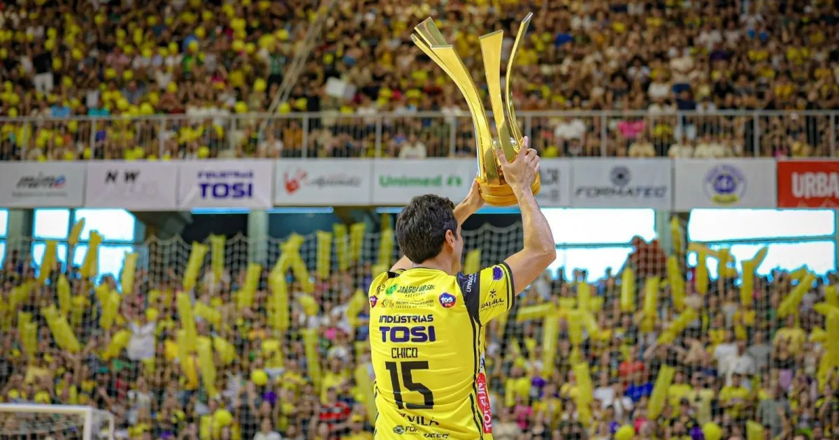 Jogador de futsal exibe troféu em estádio lotado com torcedores vestidos de amarelo
