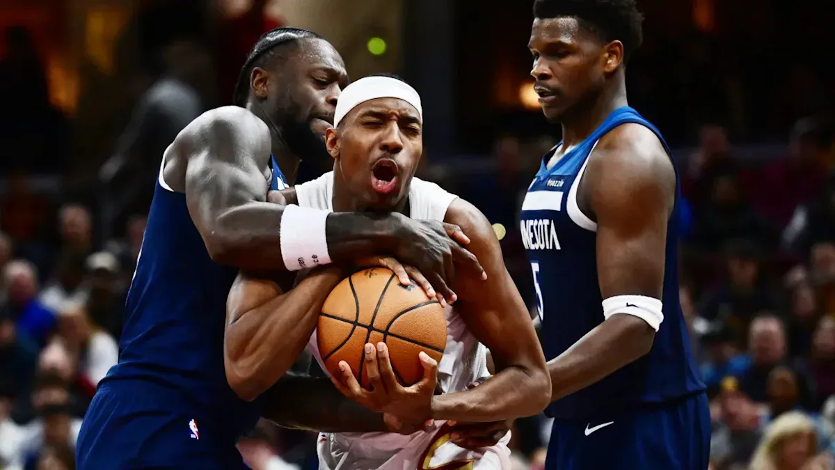 Jogador de uniforme branco disputa a posse de bola com dois adversários em uniforme azul durante partida de basquete em quadra coberta