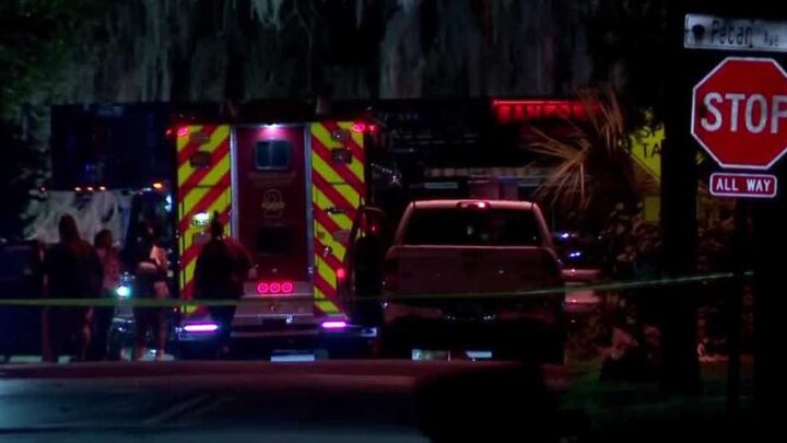 Ambulância com luzes acesas e profissionais de emergência em ação durante operação noturna em via pública, com viatura e placa de trânsito visíveis ao fundo